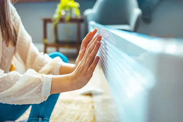 Woman in cold house putting her hands up to a heater to feel warm