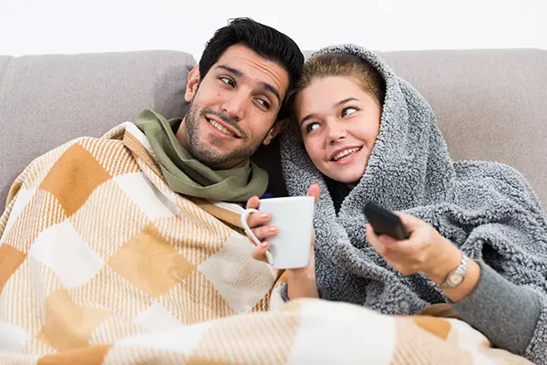 Couple sitting on the couch wrapped up in sweaters and blankets against the cold.